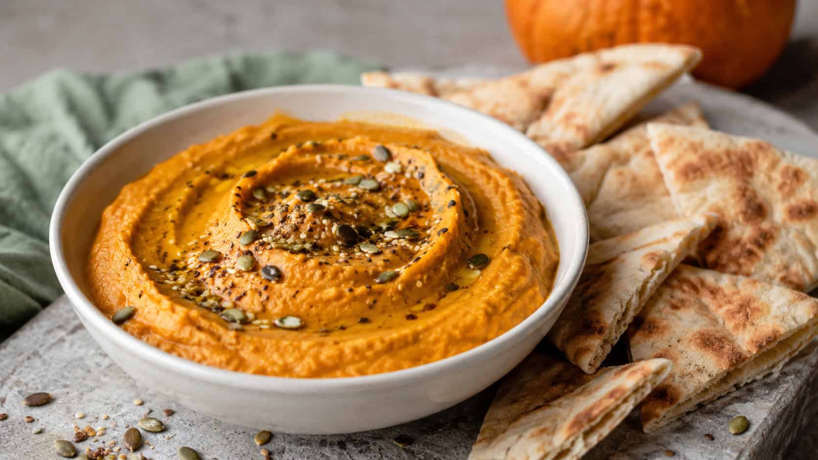 a bowl of creamy orange pumpkin hummus drizzled with olive oil beside crisp pita wedges, in a wide horizontal shot on a stone surface with a sprinkle of seeds, natural light, shallow depth of field, landscape orientation