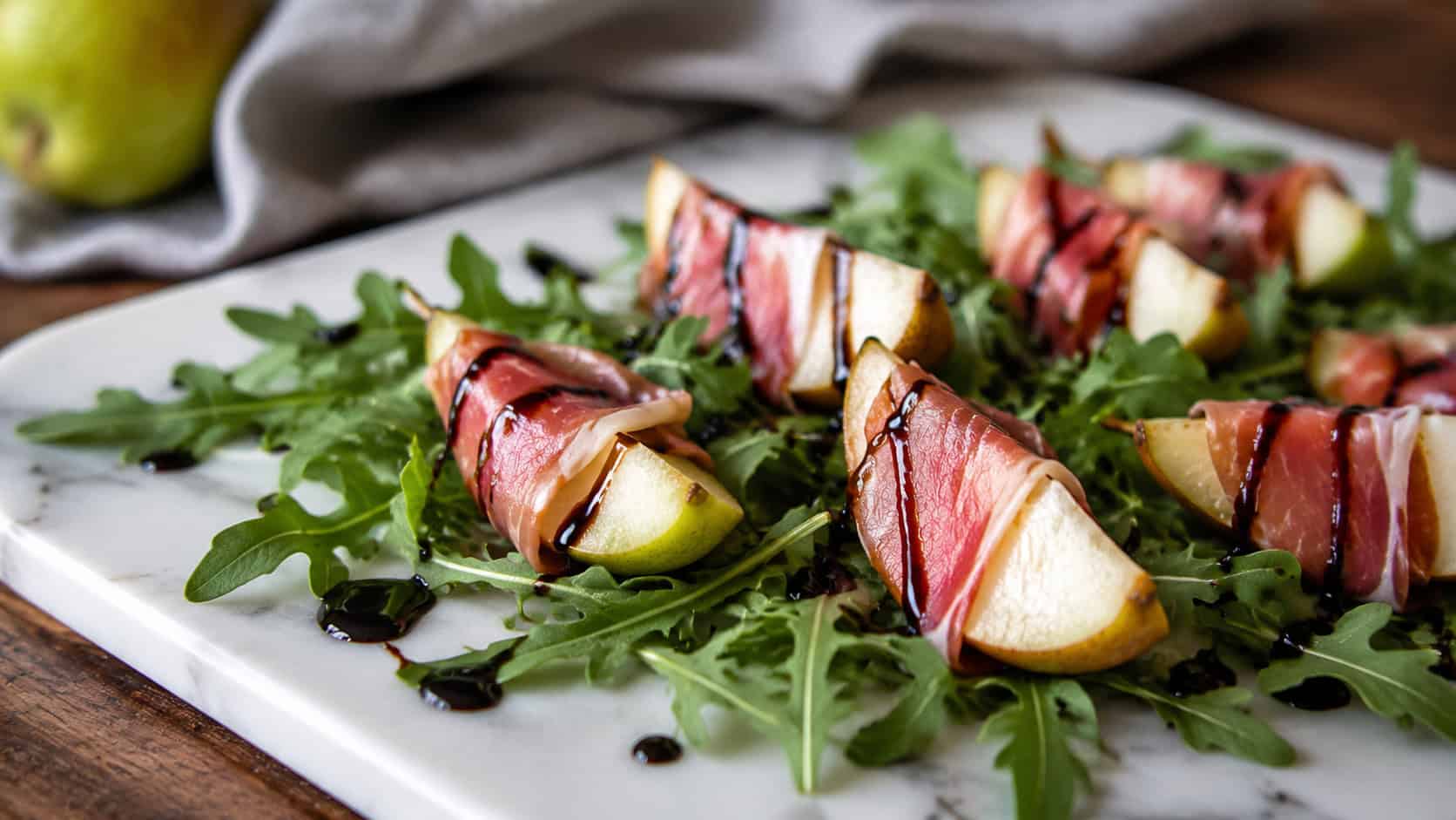 pear wedges wrapped in prosciutto drizzled with balsamic glaze over arugula, in a wide horizontal shot on a white marble platter, natural light, shallow depth of field, landscape orientation