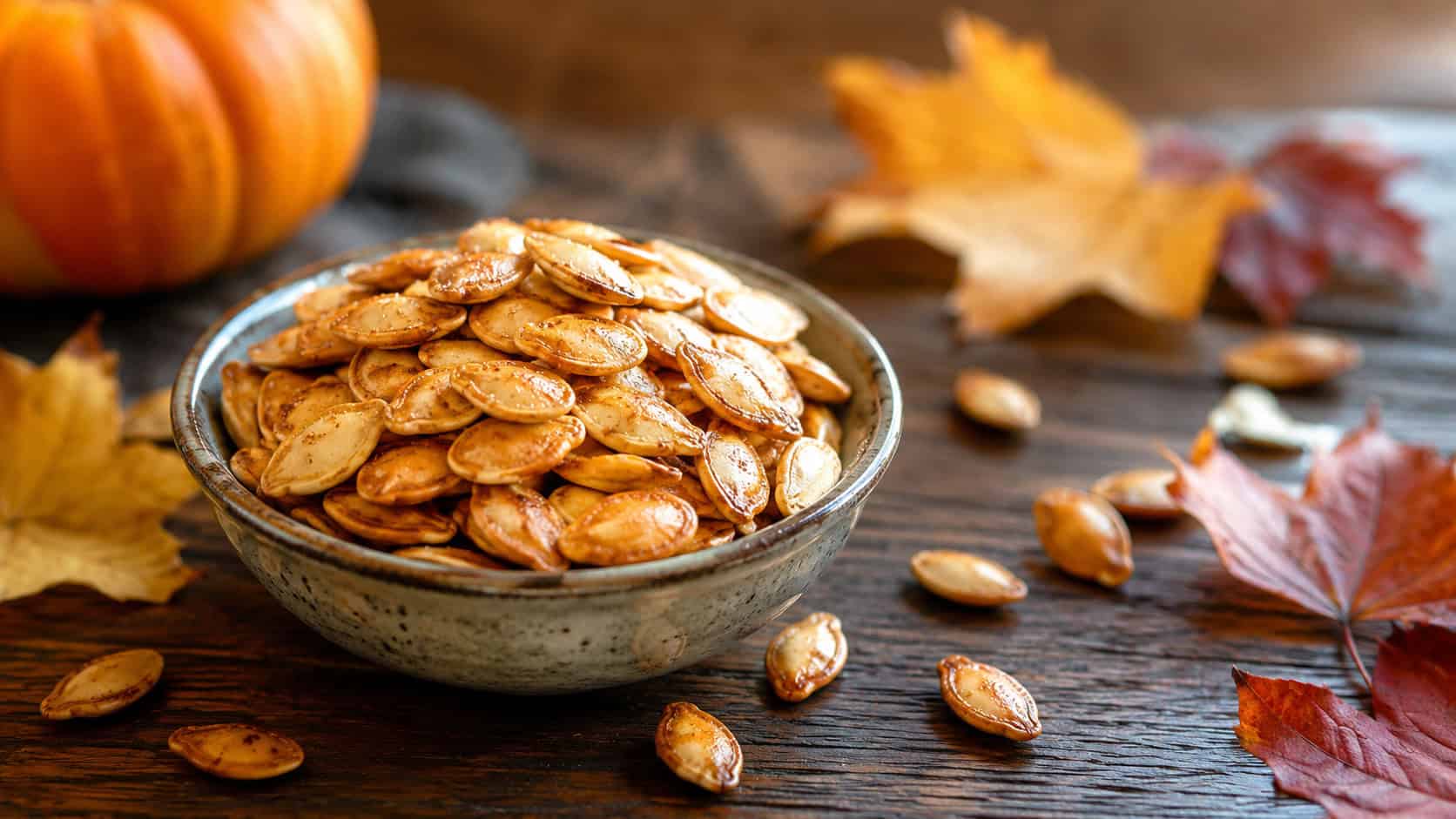 crispy golden roasted pumpkin seeds piled in a small bowl, in a wide horizontal shot on a wooden table with a scattering of seeds and autumn leaves, natural light, shallow depth of field, landscape orientation