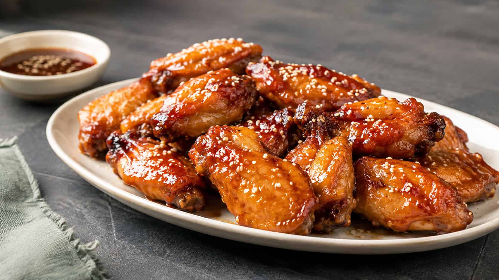 glossy honey glazed chicken wings sprinkled with sesame seeds piled on a platter, in a wide horizontal shot on a dark table with a small bowl of extra glaze, natural light, shallow depth of field, landscape orientation