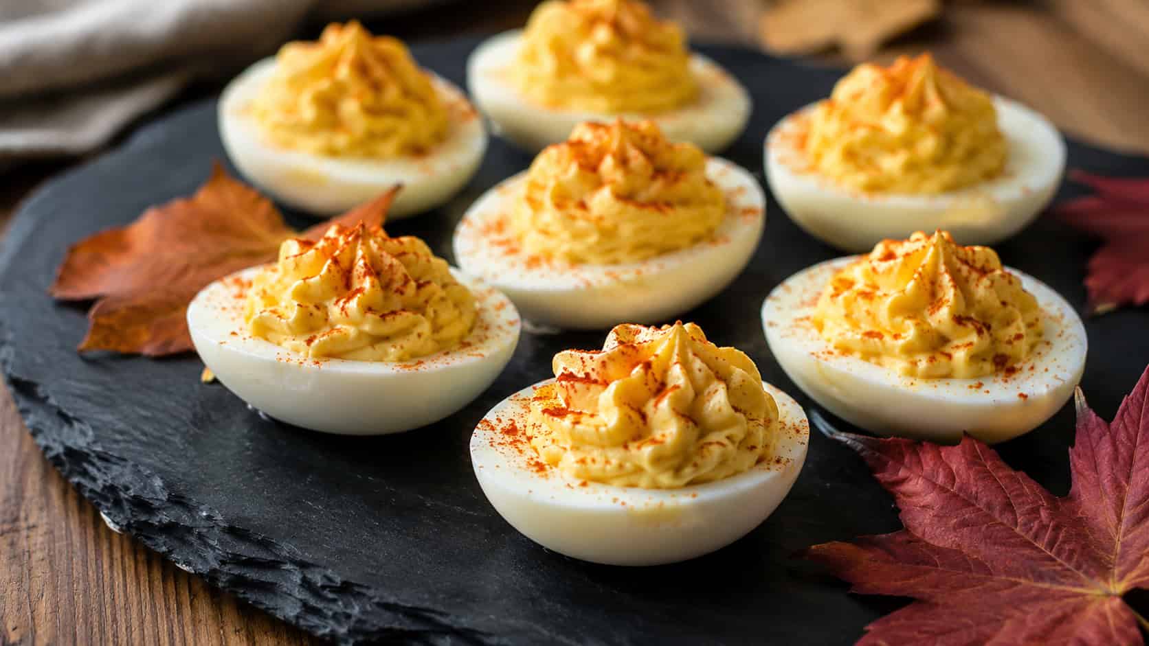 deviled eggs with piped creamy yellow filling dusted with smoked paprika, in a wide horizontal shot on a slate serving tray with a few autumn leaves, natural light, shallow depth of field, landscape orientation