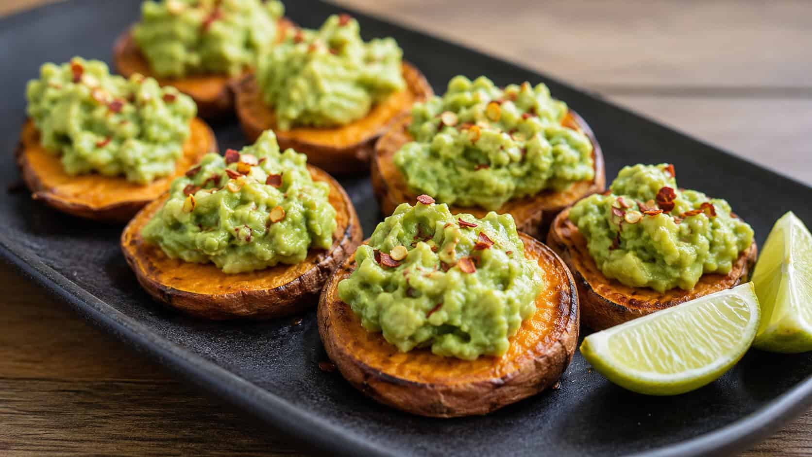 roasted sweet potato rounds topped with bright green smashed avocado and red pepper flakes, in a wide horizontal shot on a dark ceramic platter with lime wedges, natural light, shallow depth of field, landscape orientation