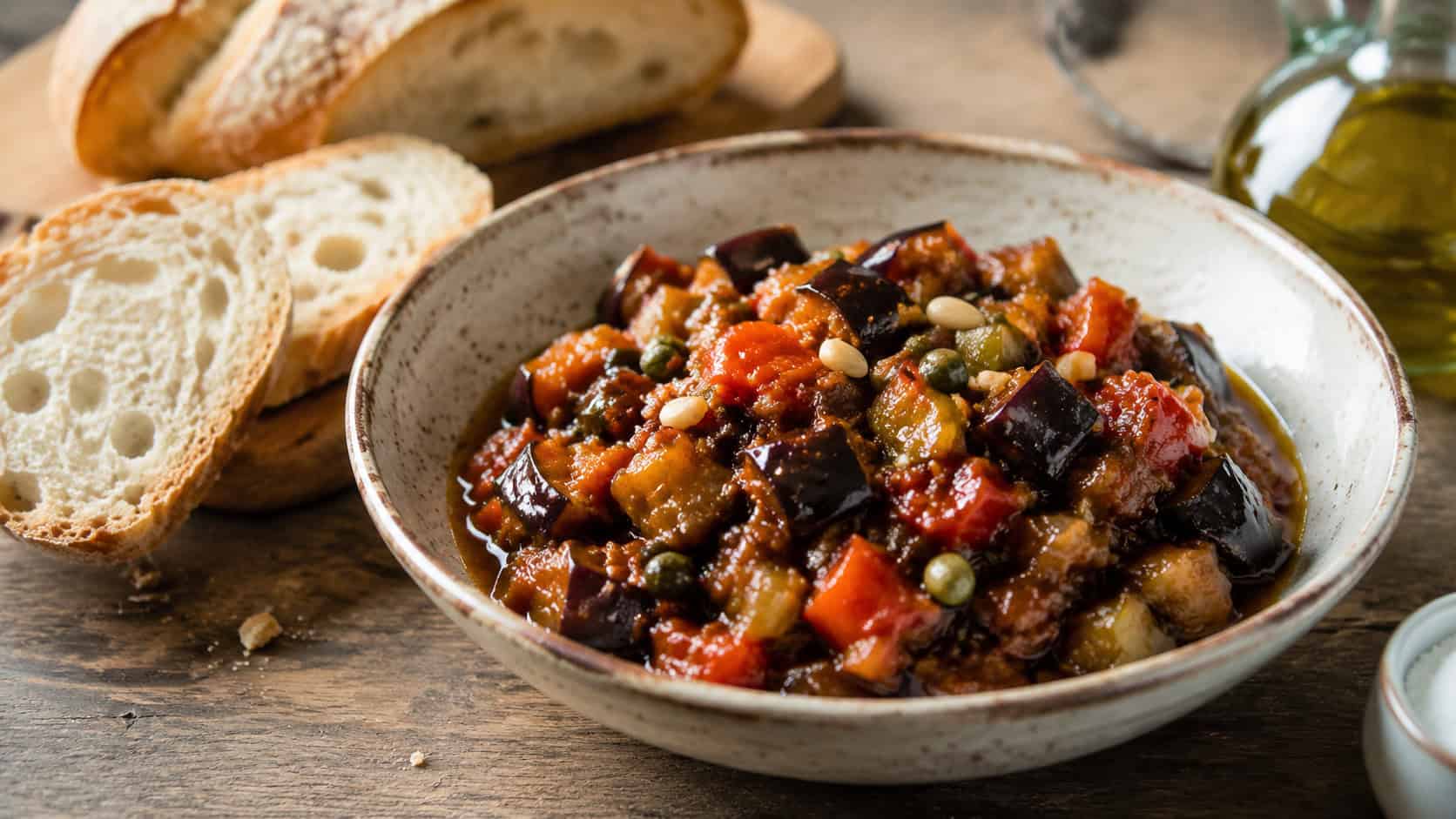 glossy eggplant caponata in a wide horizontal shot, in a rustic bowl with crusty bread and a drizzle of olive oil on a wooden table, natural light, shallow depth of field, landscape orientation