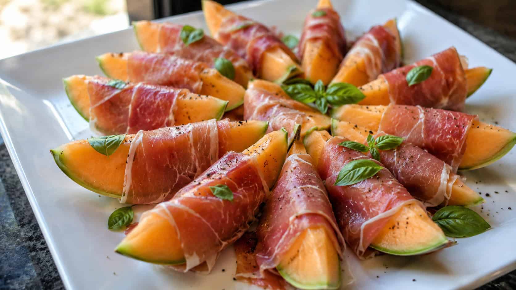 prosciutto-wrapped melon wedges in a wide horizontal shot, fanned on a white platter with cracked pepper and basil, natural light, shallow depth of field, landscape orientation