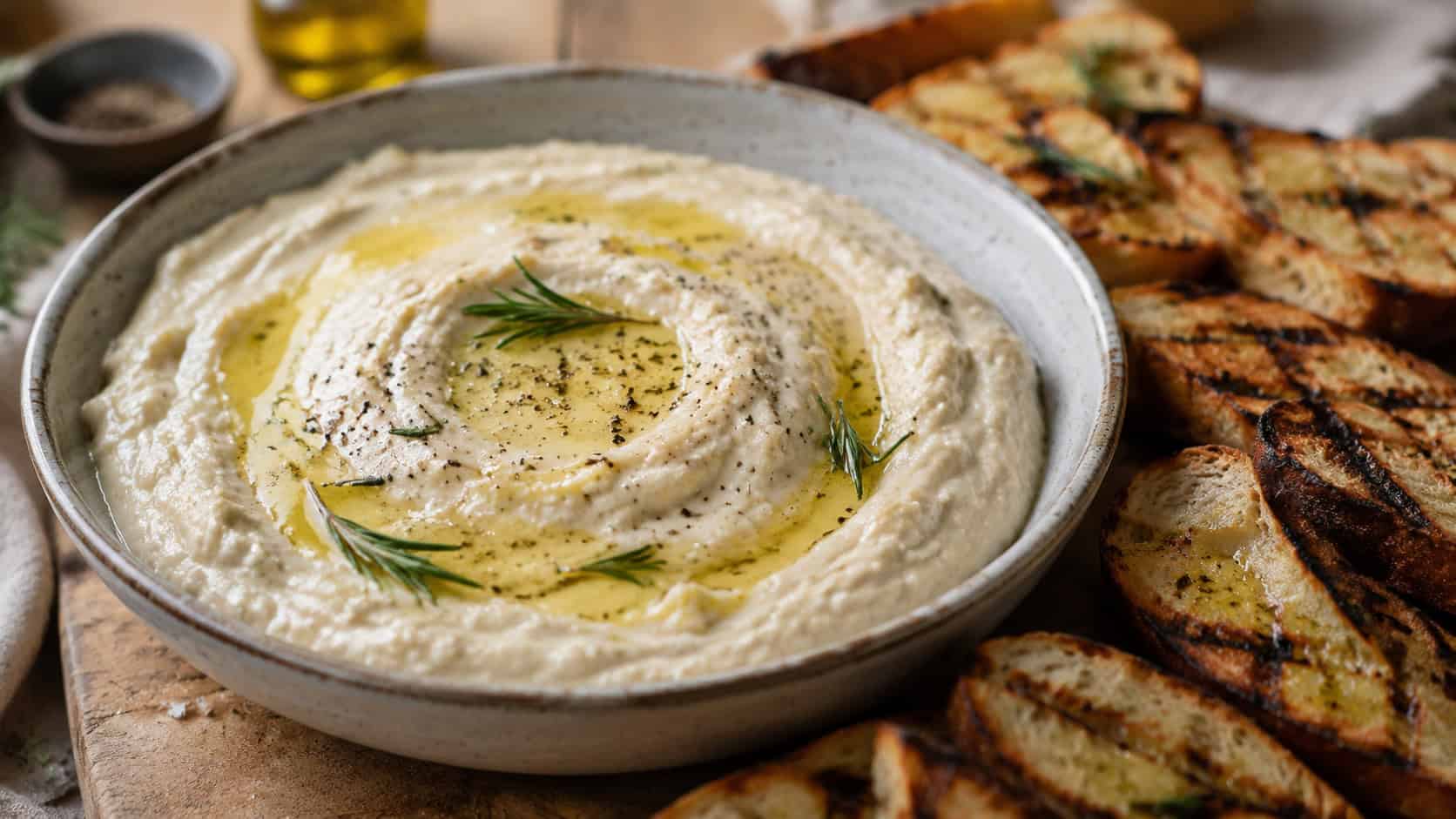 creamy white bean dip in a wide horizontal shot, in a bowl swirled with olive oil and rosemary surrounded by grilled bread, natural light, shallow depth of field, landscape orientation