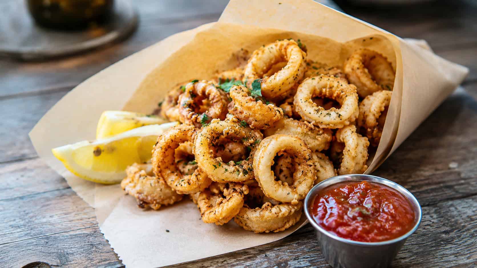 crispy fried calamari rings in a wide horizontal shot, piled in a paper cone with lemon wedges and marinara on a rustic table, natural light, shallow depth of field, landscape orientation