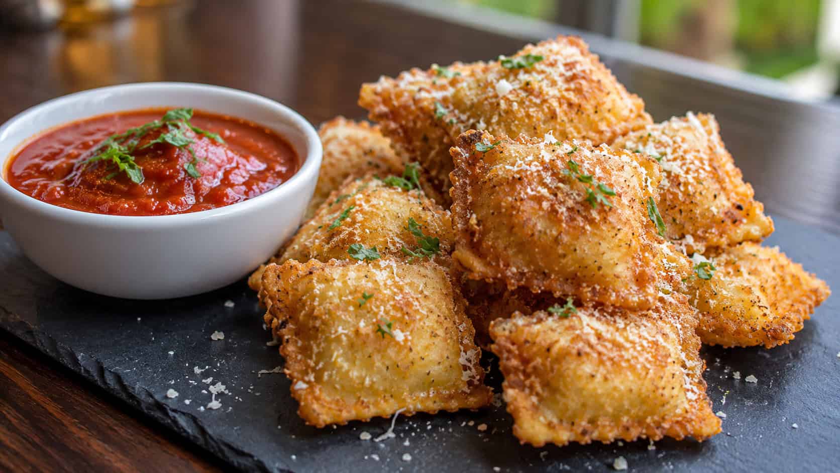 crispy fried ravioli bites in a wide horizontal shot, golden and dusted with Parmesan beside a bowl of marinara on a slate board, natural light, shallow depth of field, landscape orientation