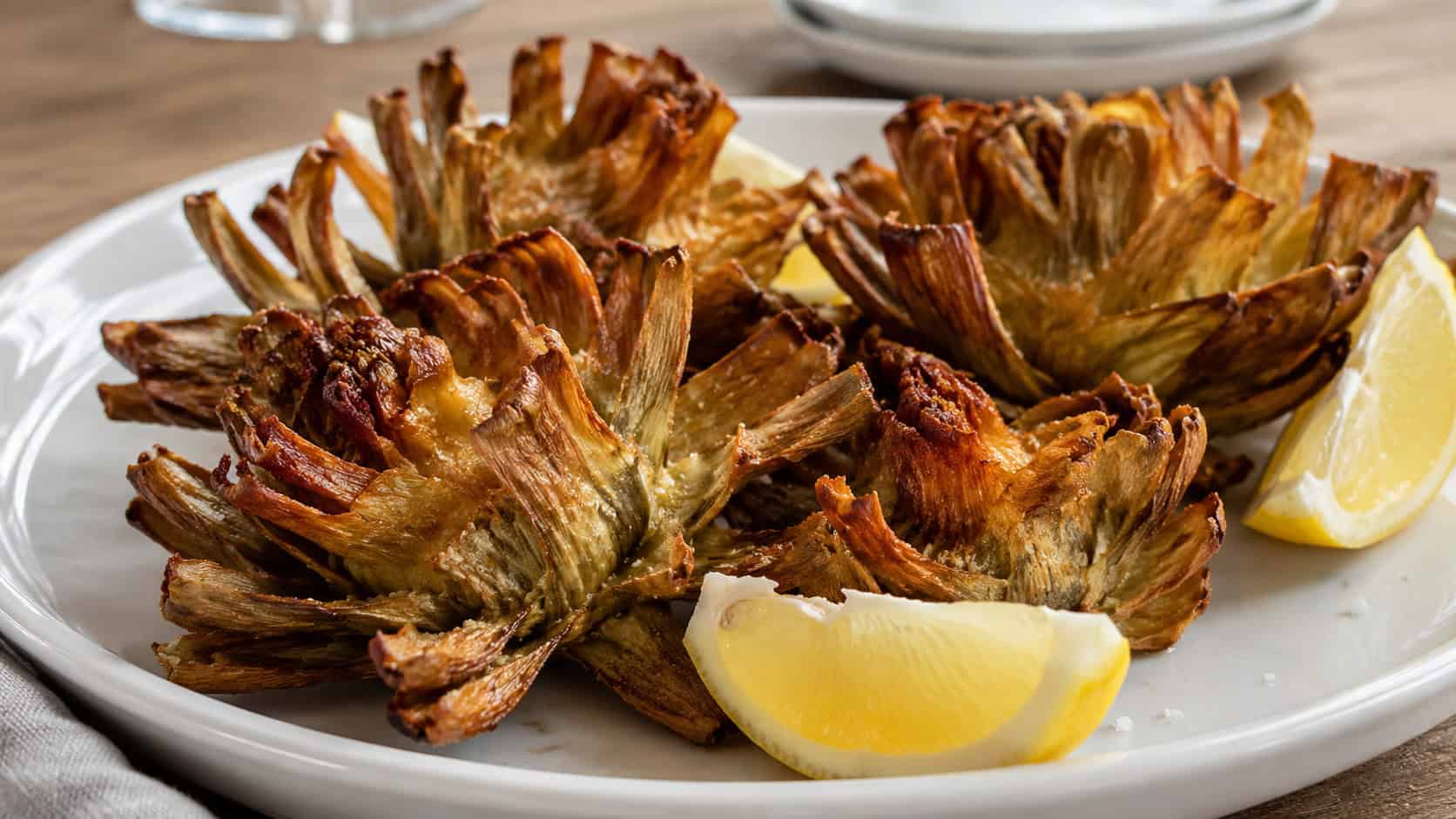 crispy fried whole artichokes in a wide horizontal shot, golden leaves splayed open like flowers on a white plate with lemon wedges, natural light, shallow depth of field, landscape orientation