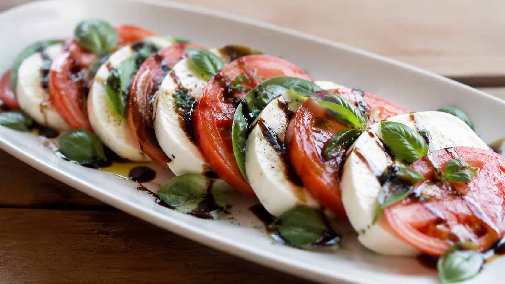 a caprese salad in a wide horizontal shot, alternating tomato and mozzarella slices with basil drizzled with balsamic on a white platter, natural light, shallow depth of field, landscape orientation