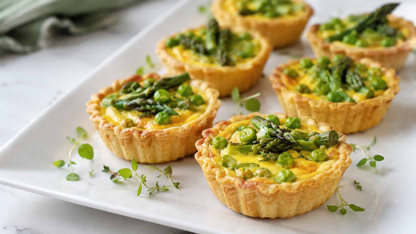 spring vegetable tartlets with peas and asparagus arranged on a wide horizontal white platter, golden custard centers, bright natural light, shallow depth of field, landscape orientation