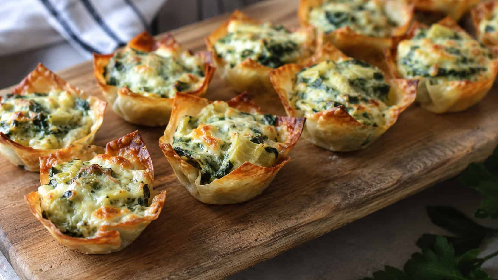 crispy wonton cups filled with bubbly spinach artichoke dip arranged on a wide horizontal serving board, golden cheesy tops, natural light, shallow depth of field, landscape orientation