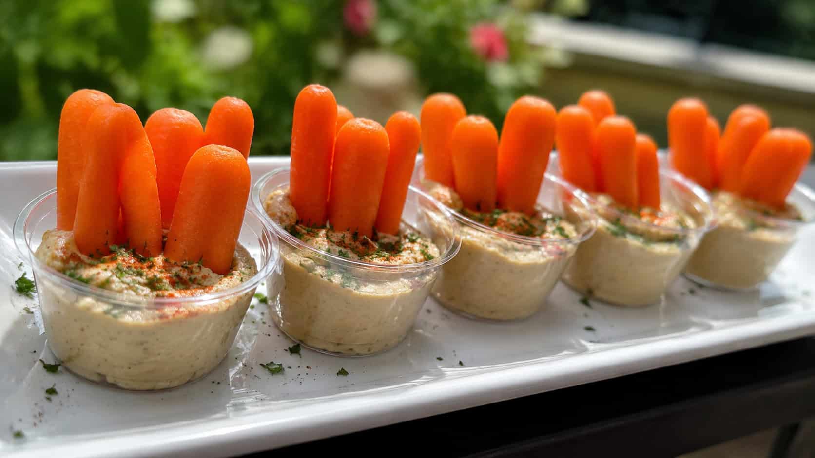 individual hummus cups with baby carrots standing upright arranged in a wide horizontal row on a white tray, paprika and parsley garnish, natural light, shallow depth of field, landscape orientation