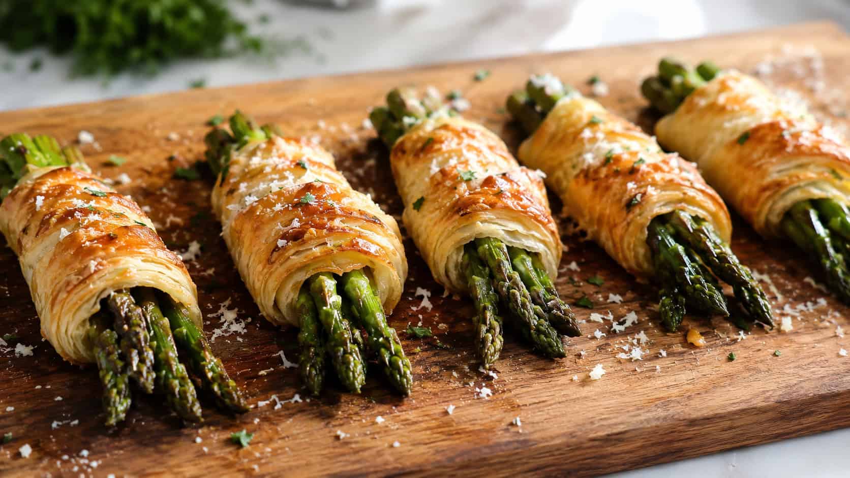 golden puff pastry asparagus bundles lined up on a wide horizontal wooden board, flaky pastry glistening, scattered Parmesan, bright natural light, shallow depth of field, landscape orientation