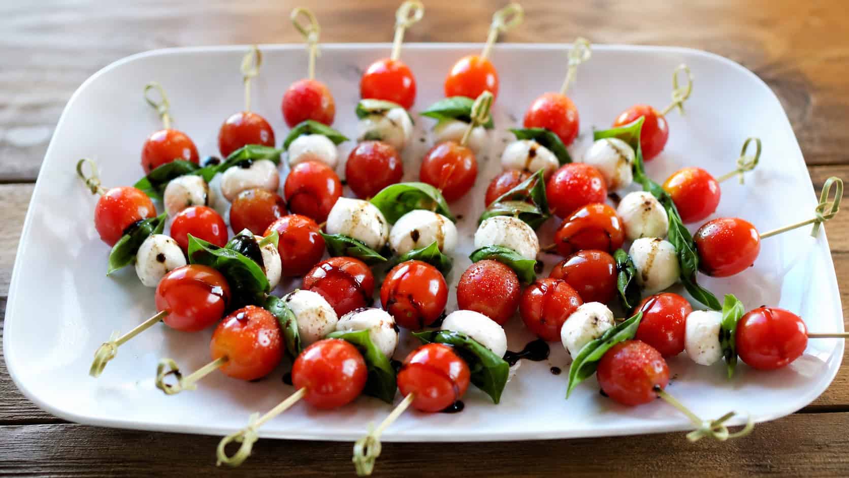 mini caprese skewers fanned out across a wide horizontal white platter, glossy cherry tomatoes and fresh basil, balsamic drizzle, natural light, shallow depth of field, landscape orientation