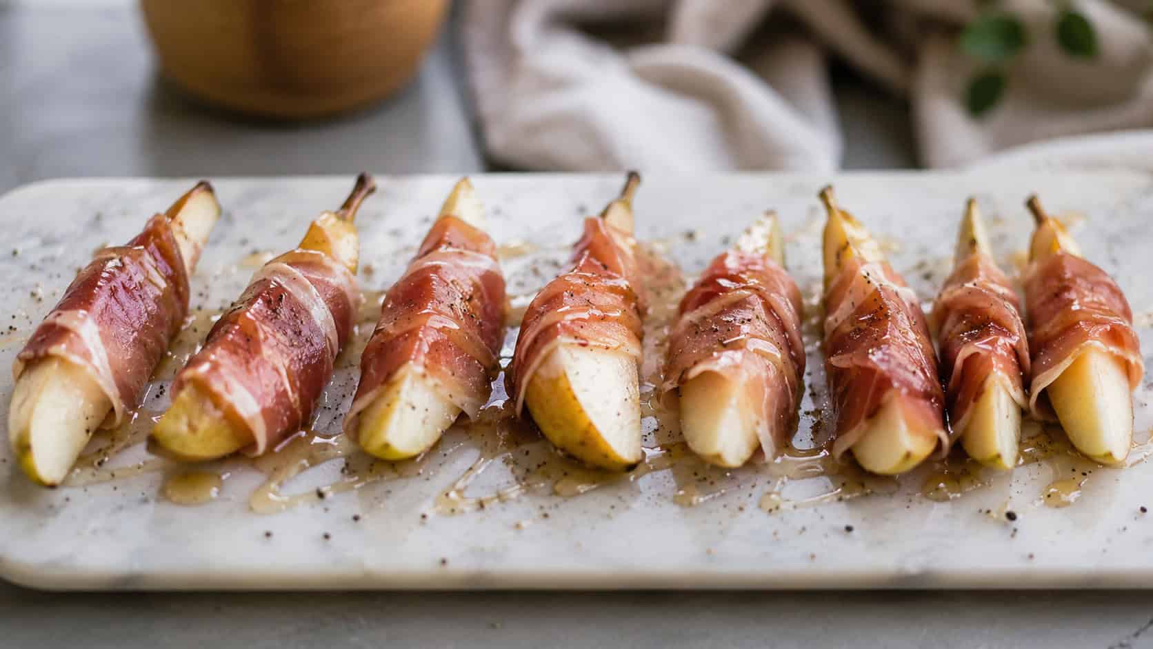 prosciutto-wrapped pear wedges arranged in a wide horizontal line on a marble board, glistening honey drizzle and cracked pepper, soft natural light, shallow depth of field, landscape orientation