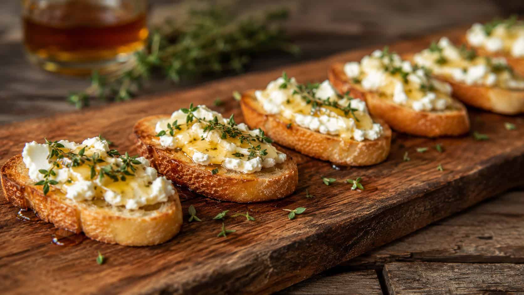 goat cheese crostini drizzled with honey and sprinkled with thyme, lined up on a wide horizontal rustic wooden board, warm natural light, shallow depth of field, landscape orientation