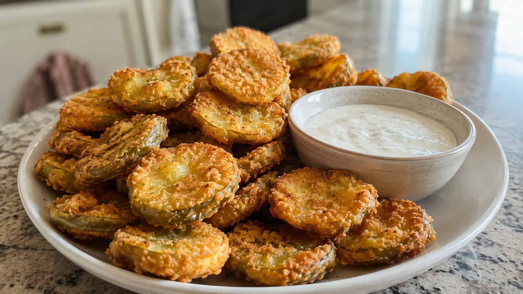 golden crispy fried pickle chips piled on a wide plate with a bowl of ranch dip, set on a counter, natural light, shallow depth of field, landscape orientation