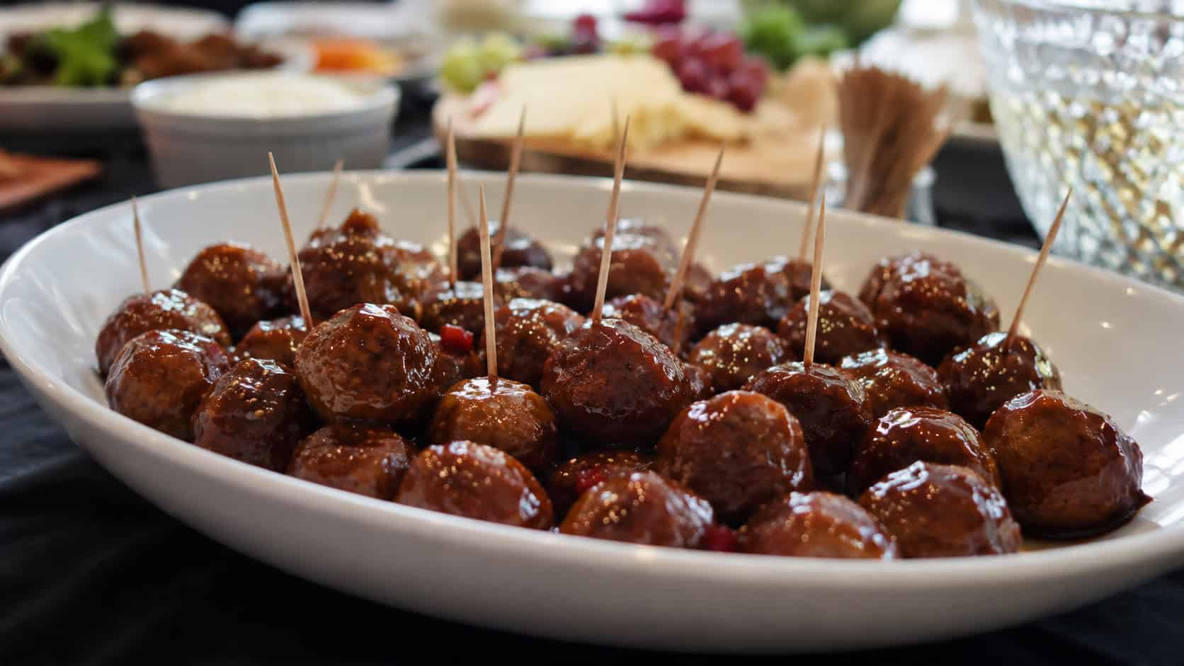 glossy sweet and spicy glazed meatballs in a wide serving dish with toothpicks, set on a party table, natural light, shallow depth of field, landscape orientation