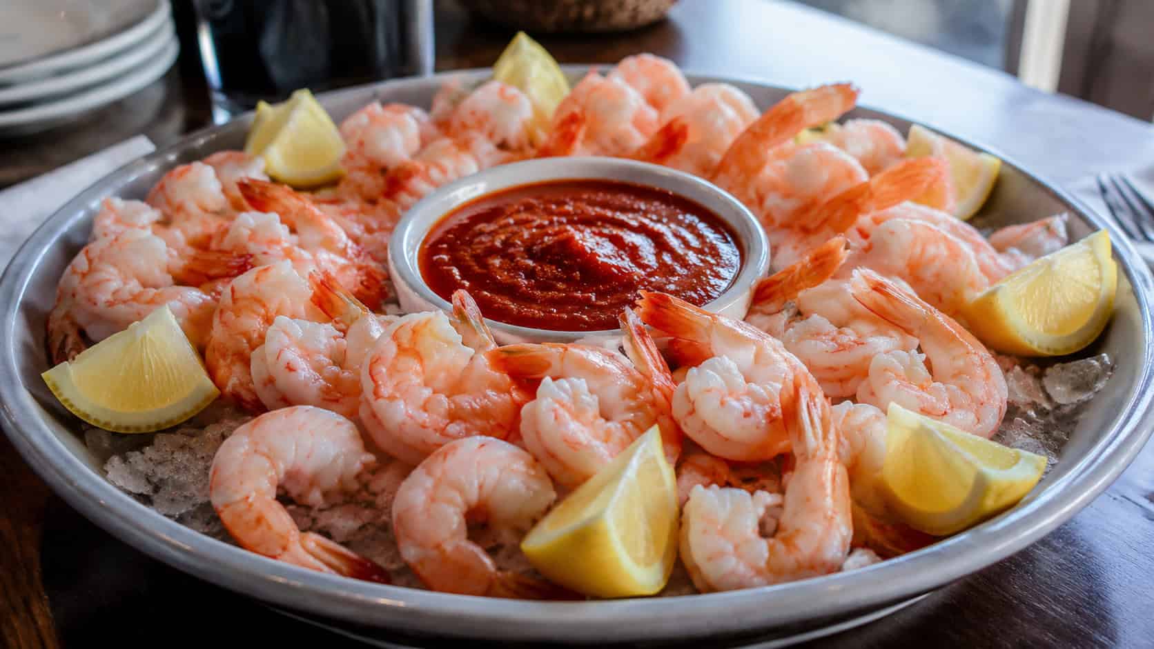 chilled shrimp cocktail arranged around a bowl of red cocktail sauce on a wide platter with lemon wedges, set on a table, natural light, shallow depth of field, landscape orientation