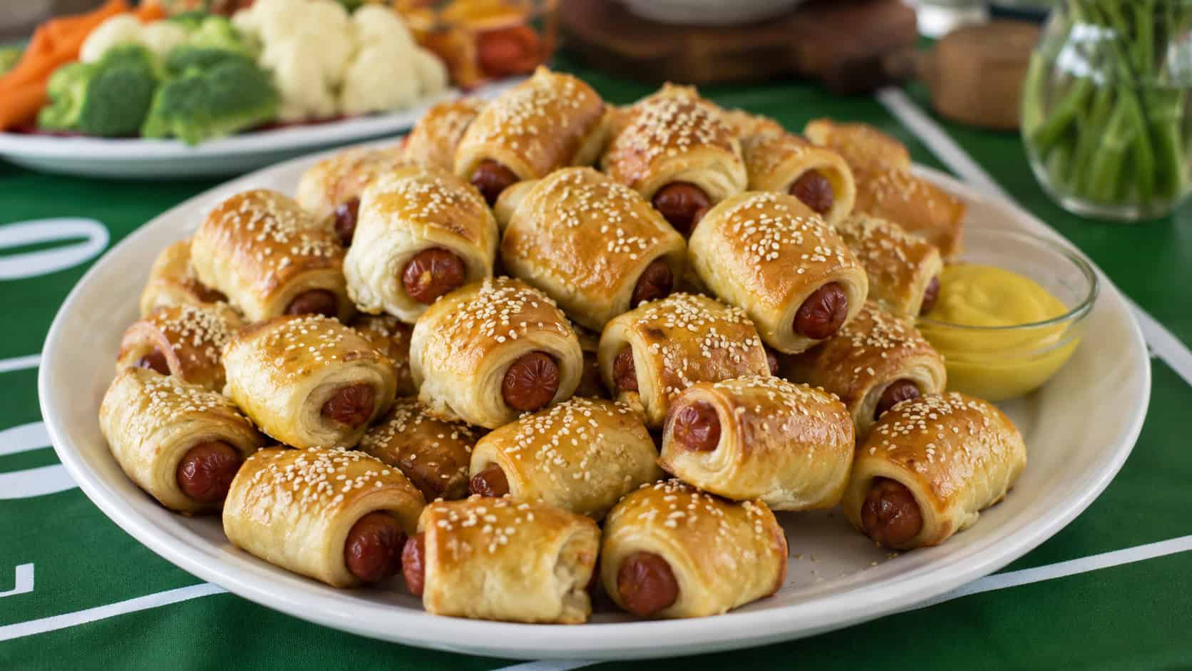 golden pigs in a blanket sprinkled with sesame seeds piled on a wide platter with mustard, set on a party table, natural light, shallow depth of field, landscape orientation