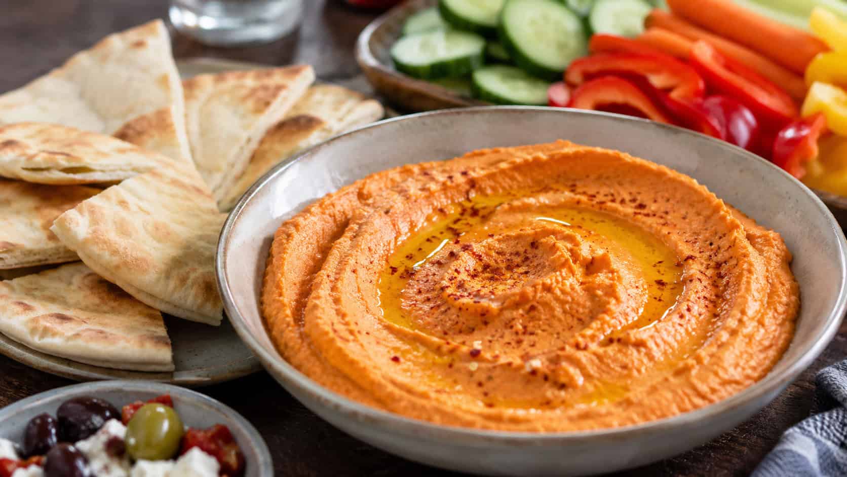 creamy roasted red pepper hummus in a wide bowl with an olive oil swirl, surrounded by pita wedges and vegetables on a table, natural light, shallow depth of field, landscape orientation
