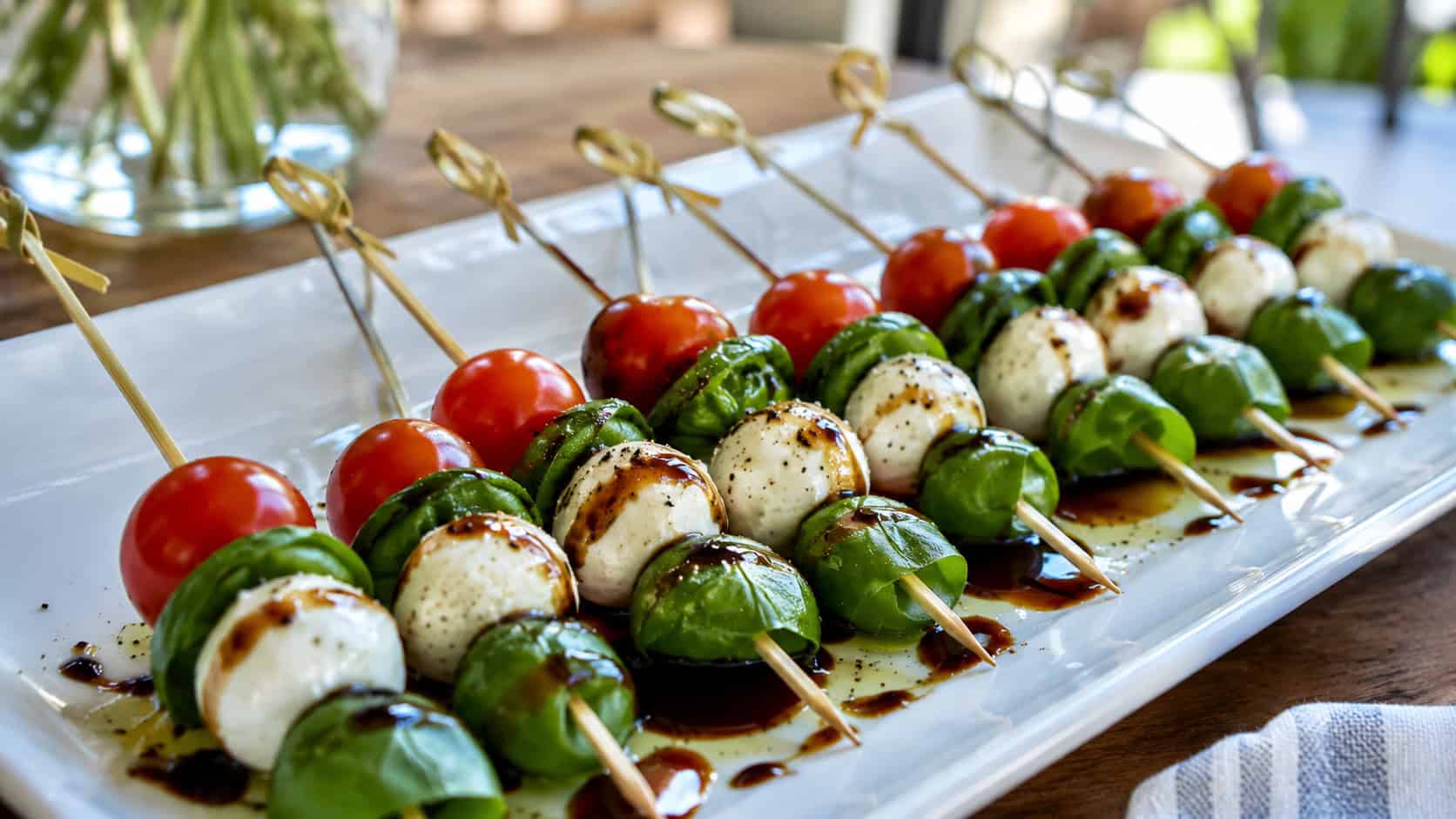 caprese salad skewers with cherry tomatoes, mozzarella, and basil drizzled with balsamic, lined up on a wide white platter on a table, natural light, shallow depth of field, landscape orientation