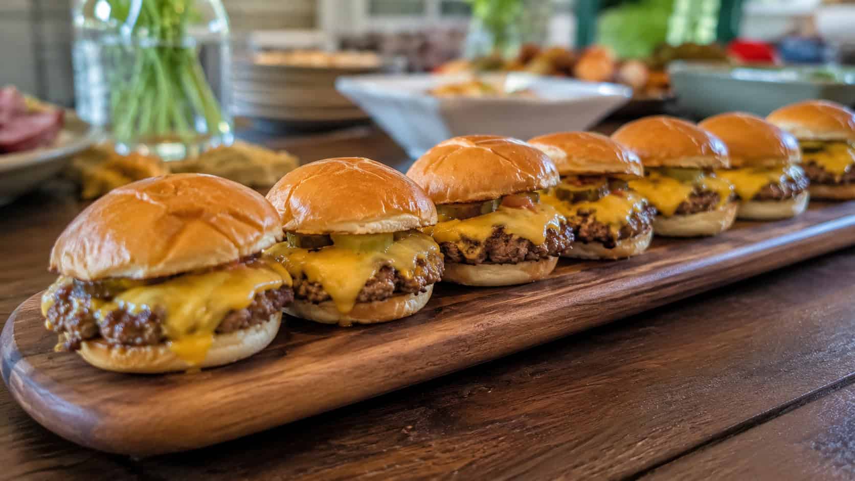 mini cheeseburger sliders with melted cheese on soft buns, arranged in a wide row on a serving board on a party table, natural light, shallow depth of field, landscape orientation