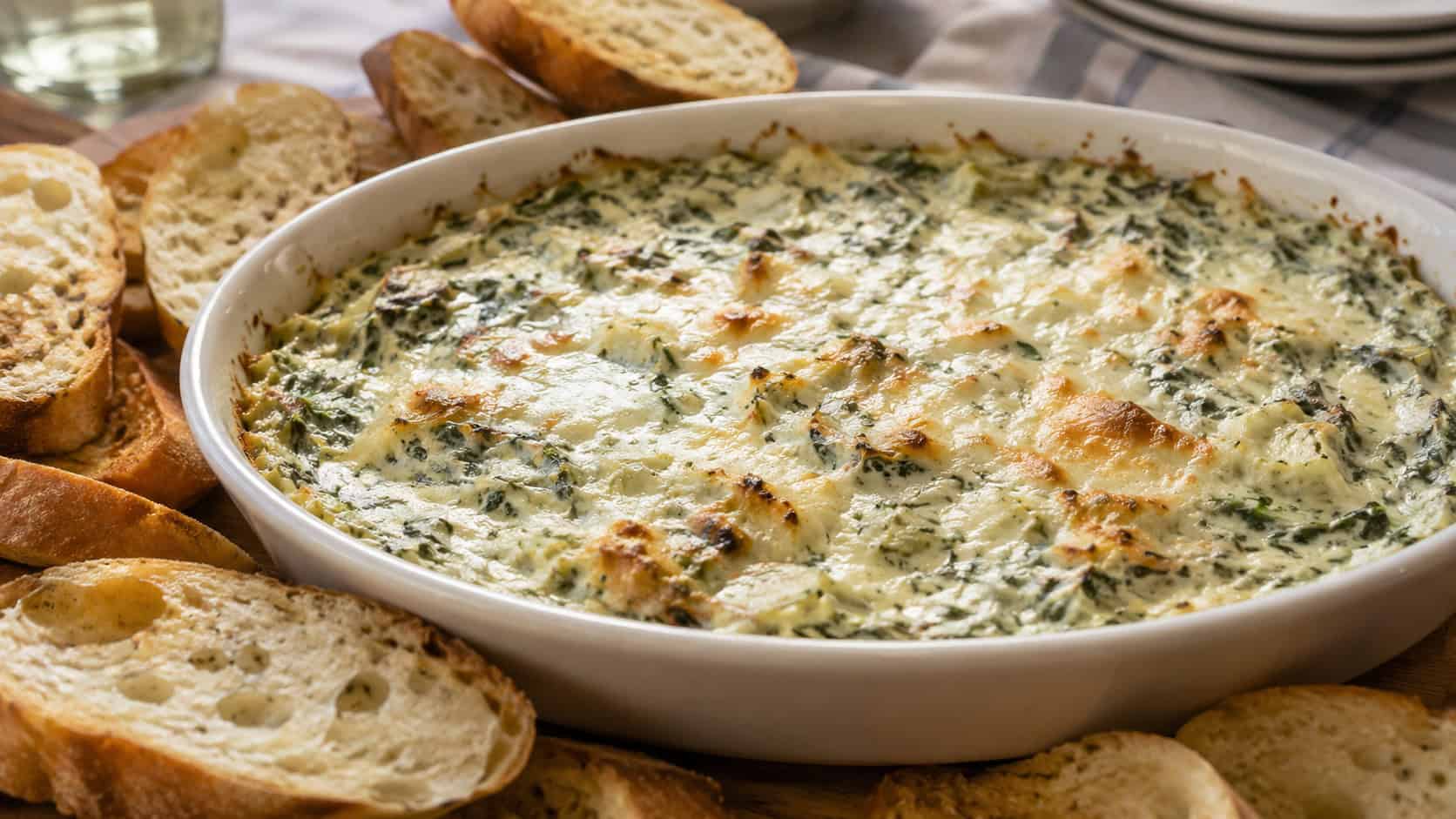 golden bubbly spinach artichoke dip in a wide oval dish with crusty bread slices around it on a table, natural light, shallow depth of field, landscape orientation