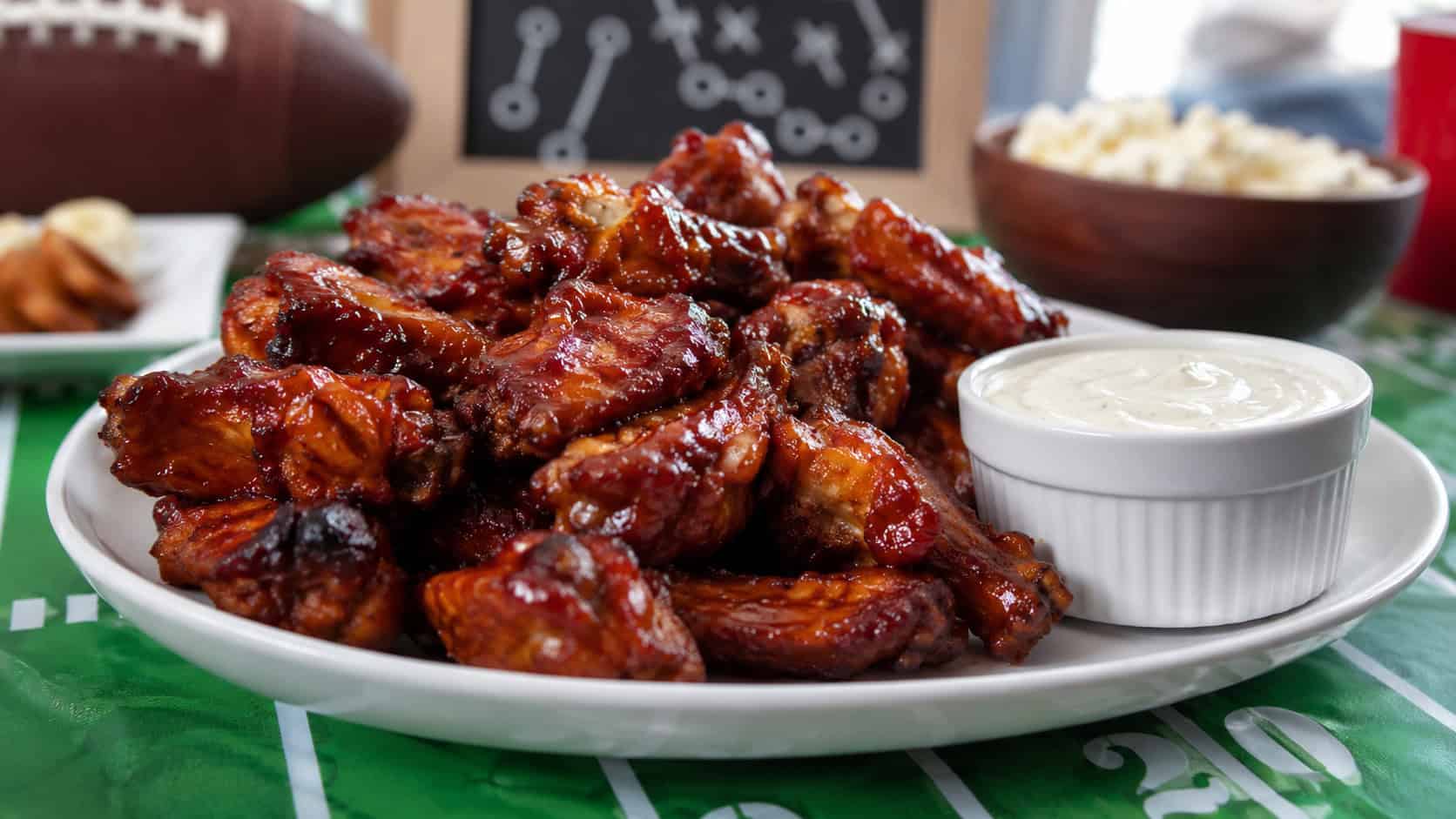 glossy barbecue chicken wings piled on a wide platter with a dipping bowl on the side, set on a game-day table, natural light, shallow depth of field, landscape orientation