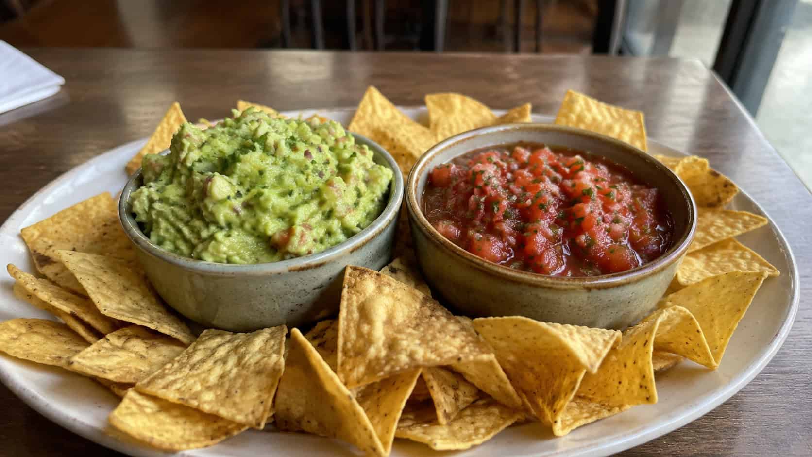 fresh guacamole and red salsa in two bowls on a wide platter surrounded by tortilla chips on a table, natural light, shallow depth of field, landscape orientation