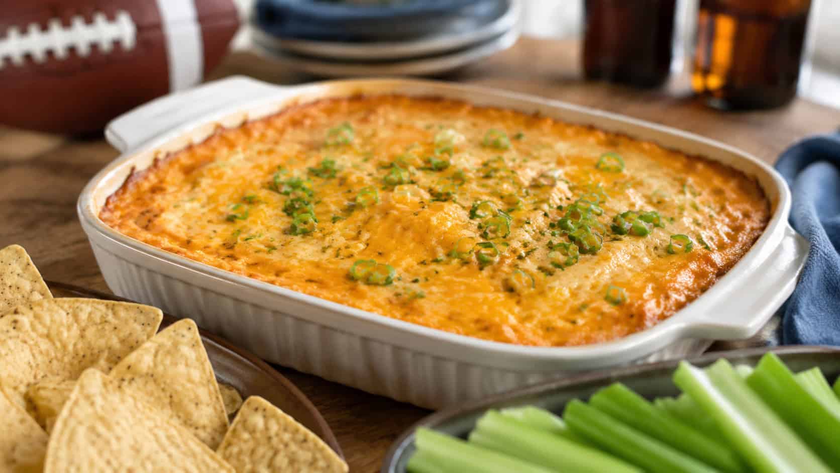 a bubbling buffalo chicken dip in a wide baking dish topped with melted cheddar and green onions, surrounded by tortilla chips and celery, in a wide horizontal shot, on a game day table, natural light, shallow depth of field, landscape orientation