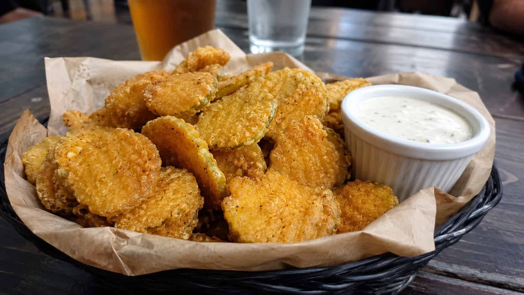 a wide basket of crispy golden fried pickle chips with a ramekin of ranch, in a wide horizontal shot, on a casual pub-style table, natural light, shallow depth of field, landscape orientation