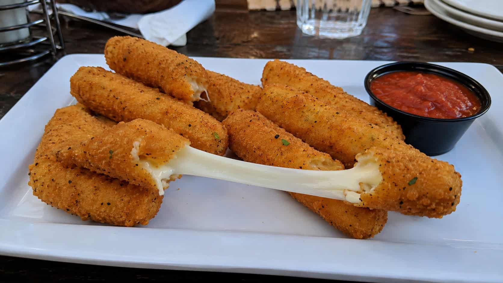 golden mozzarella sticks with one broken open showing a long cheese pull, on a wide plate with marinara, in a wide horizontal shot, on a casual table, natural light, shallow depth of field, landscape orientation
