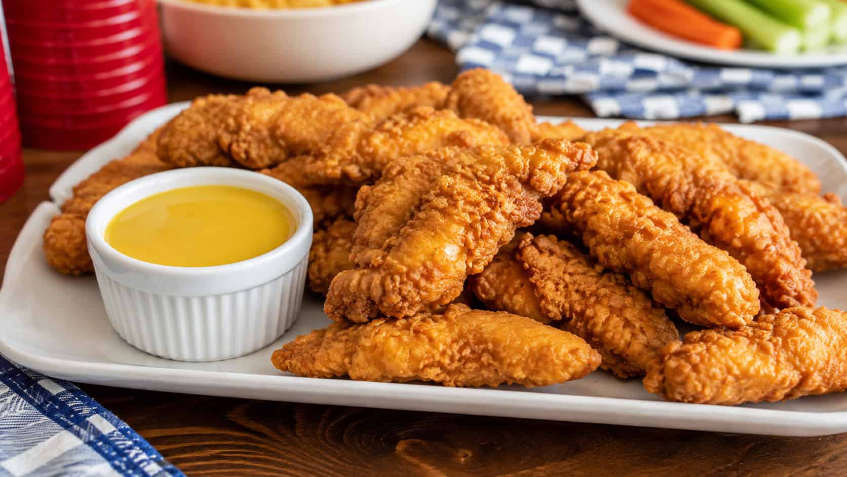 golden crispy chicken tenders with a ramekin of honey mustard on a wide platter, in a wide horizontal shot, on a casual party table, natural light, shallow depth of field, landscape orientation