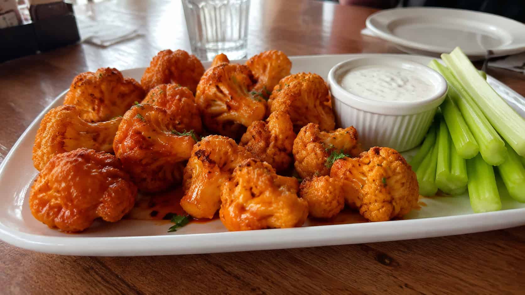 crispy buffalo cauliflower bites glistening in orange sauce on a wide platter with a ramekin of ranch and celery, in a wide horizontal shot, on a casual table, natural light, shallow depth of field, landscape orientation