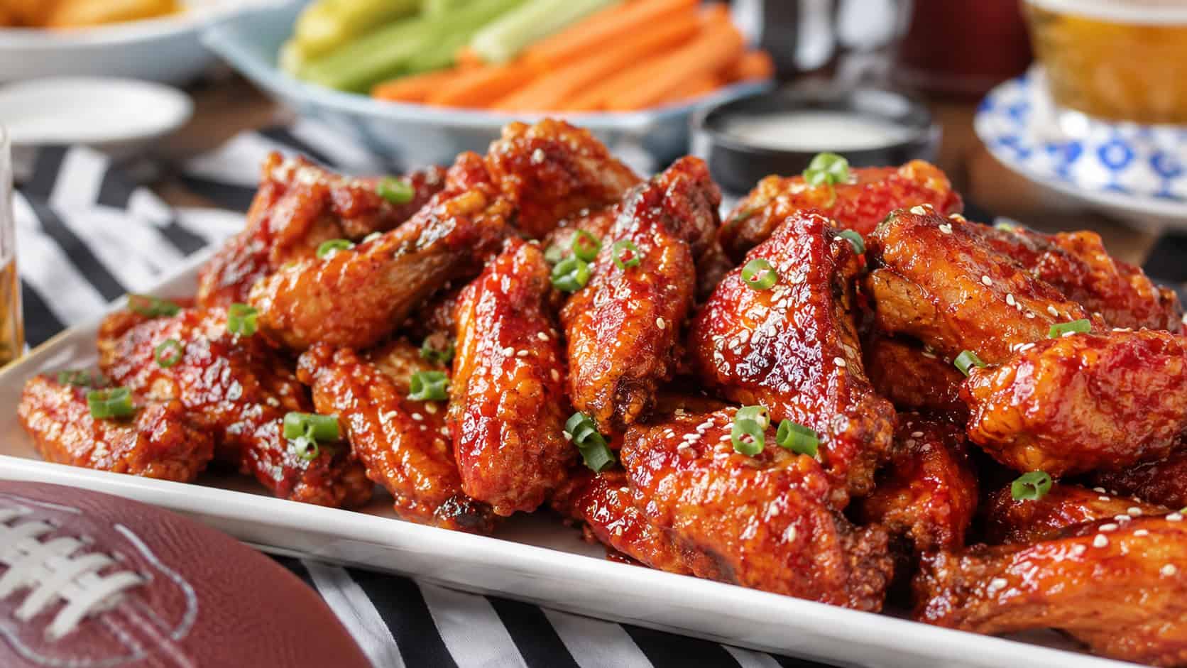 glossy spicy sriracha chicken wings glistening with red glaze and sesame seeds piled on a wide platter, in a wide horizontal shot, on a casual game day table, natural light, shallow depth of field, landscape orientation