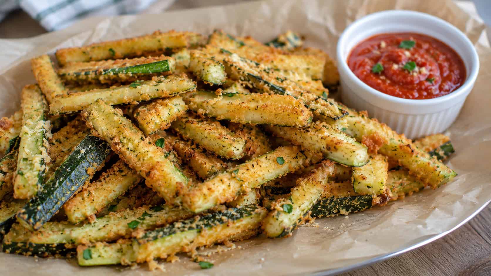 a wide pile of crispy golden parmesan zucchini fries with a ramekin of marinara, in a wide horizontal shot, on a parchment-lined platter, natural light, shallow depth of field, landscape orientation
