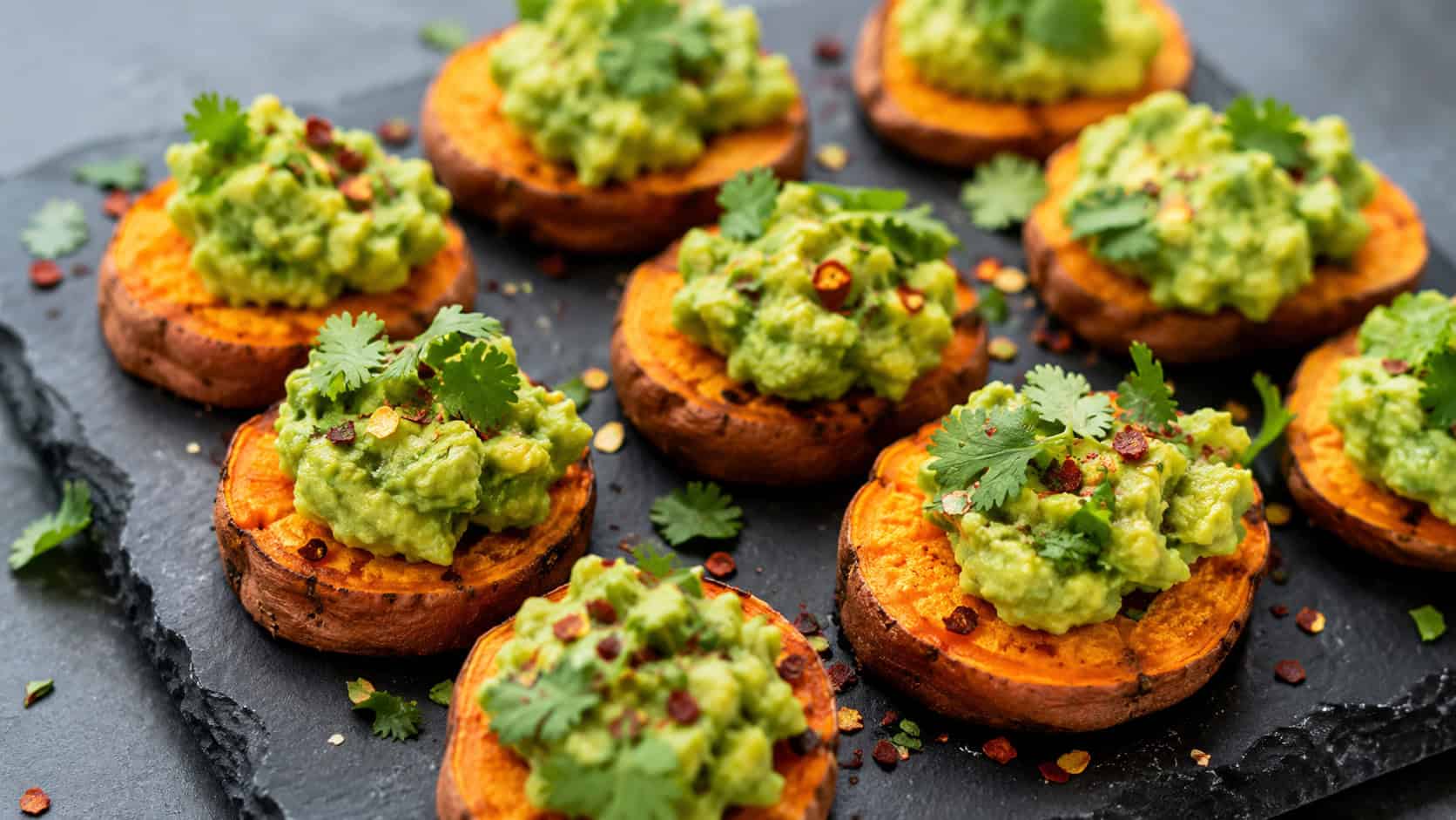 roasted sweet potato rounds topped with mashed avocado and cilantro on a slate board, wide horizontal shot, vibrant orange and green, red pepper flakes, natural light, shallow depth of field, landscape orientation