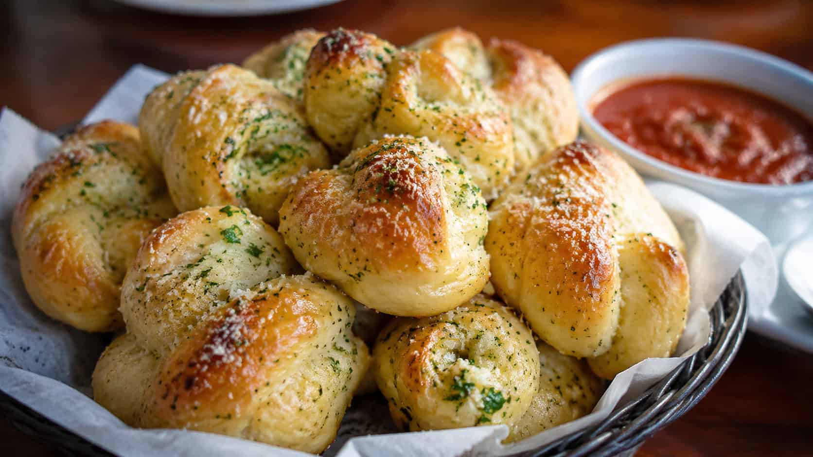 golden parmesan garlic knots piled in a basket with parsley garnish and marinara on the side, wide horizontal shot, glossy buttery tops, warm natural light, shallow depth of field, landscape orientation