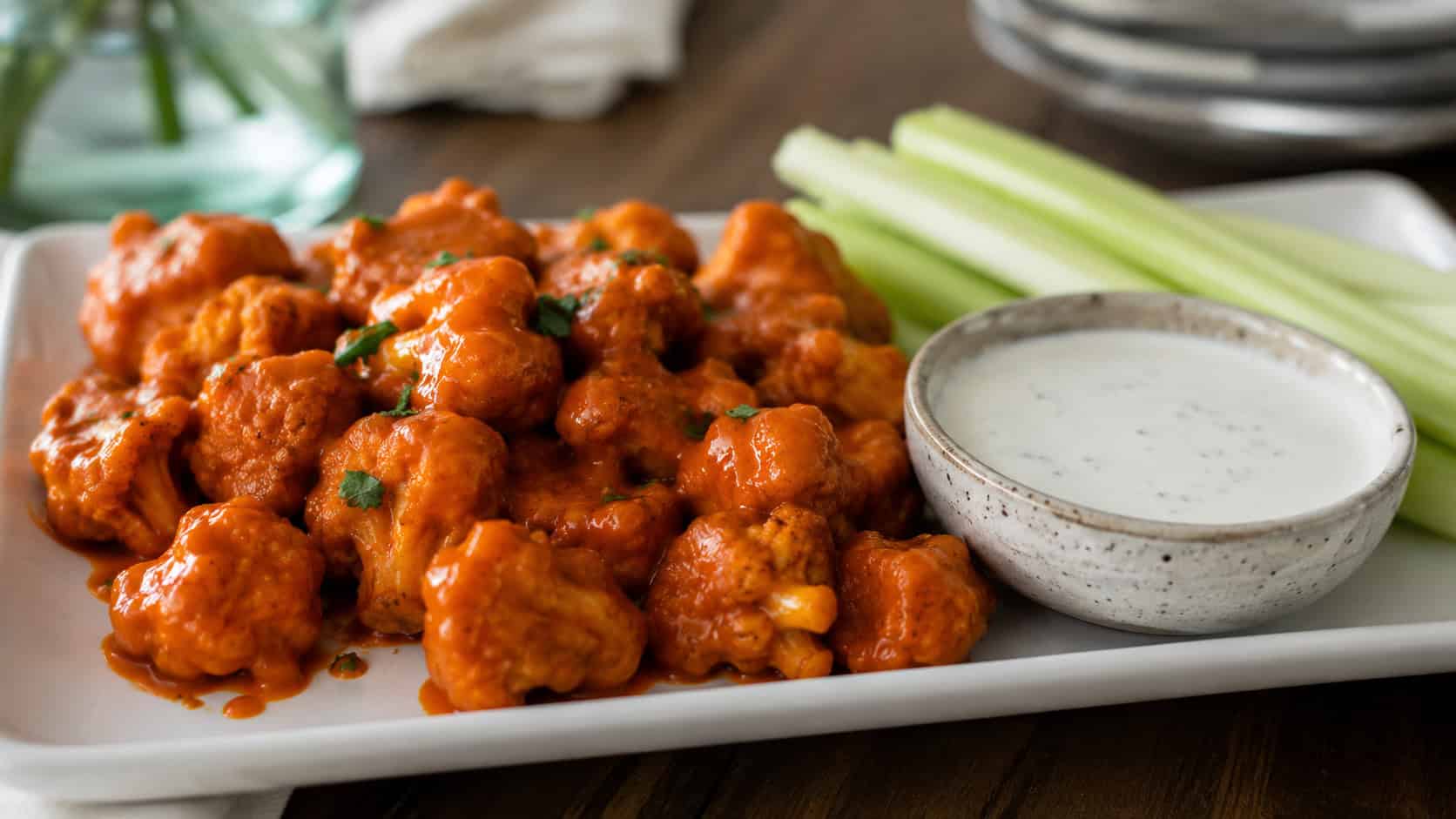 buffalo cauliflower bites arranged wide in landscape orientation on a platter, glossy orange-glazed florets beside a bowl of ranch with celery sticks, natural light, shallow depth of field, soft background blur