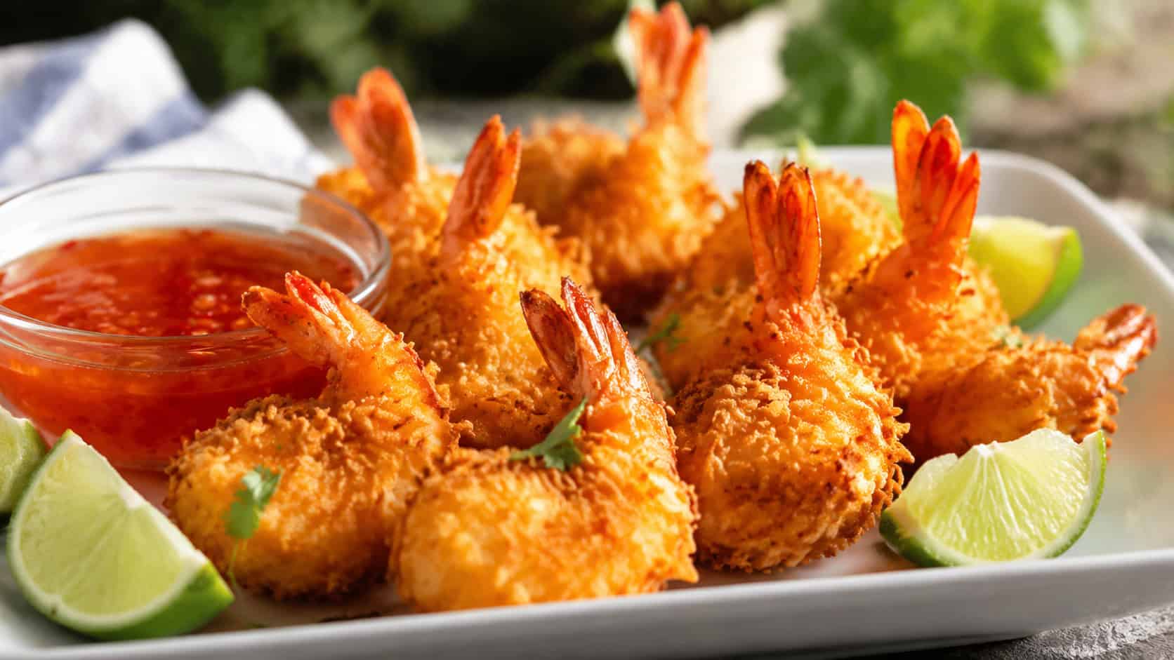 golden coconut shrimp arranged wide in landscape orientation on a platter, crispy coconut-crusted shrimp with tails up beside a bowl of sweet chili sauce, lime wedges, natural light, shallow depth of field, soft background blur