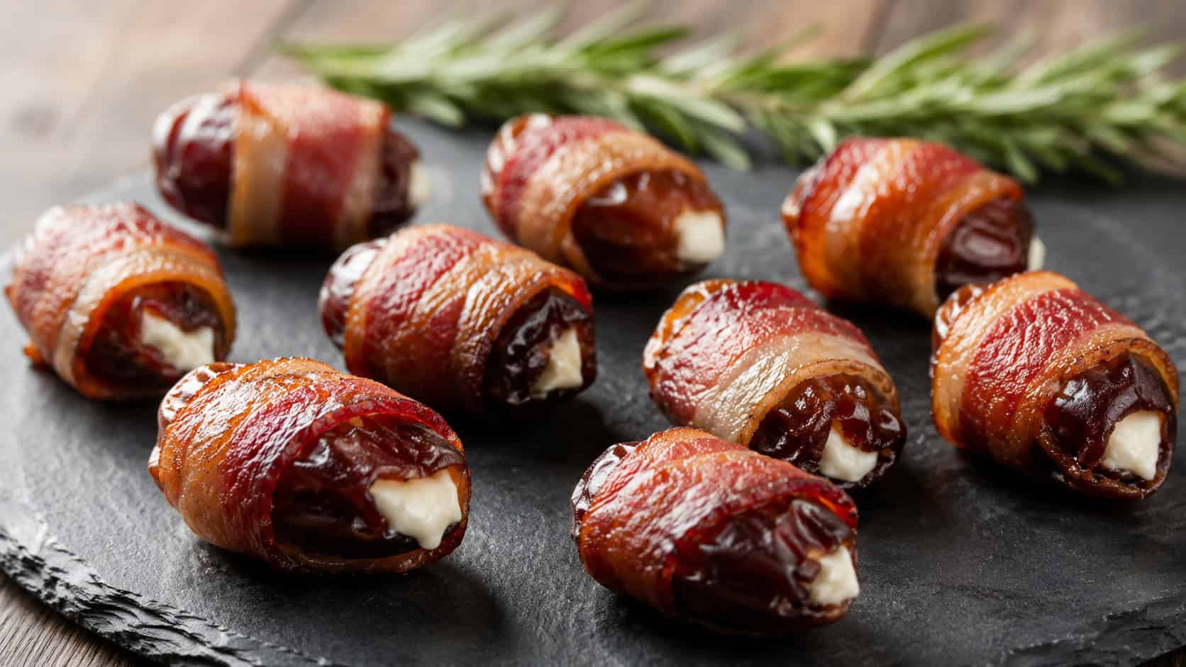bacon-wrapped dates arranged wide in landscape orientation on a slate serving board, glossy crisp bacon with peeks of creamy filling, natural light, shallow depth of field, a sprig of rosemary blurred in the background