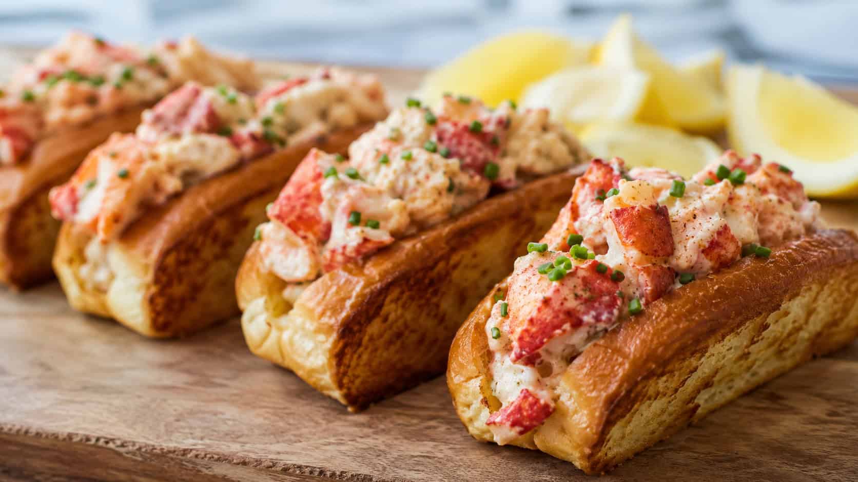 mini lobster rolls arranged wide in landscape orientation on a wooden board, golden toasted buns overflowing with chunky lobster salad and chives, natural light, shallow depth of field, lemon wedges blurred in the background