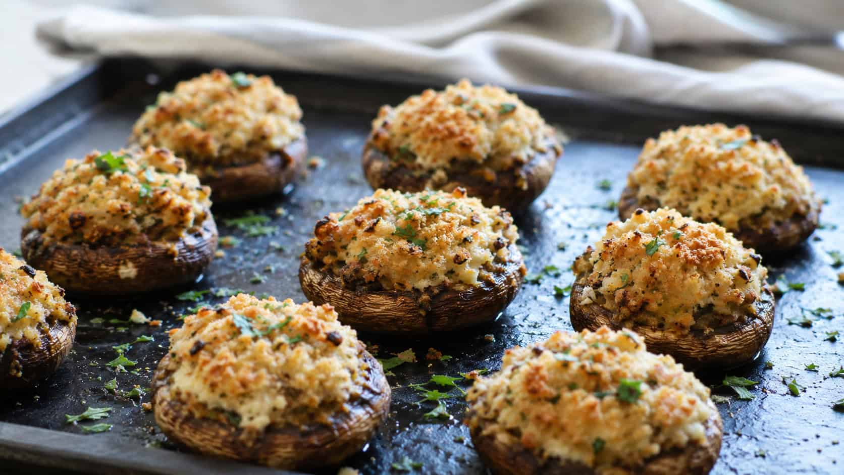 stuffed mushrooms arranged wide in landscape orientation on a dark baking tray, golden parmesan breadcrumb tops sprinkled with parsley, natural light, shallow depth of field, a linen napkin blurred in the background