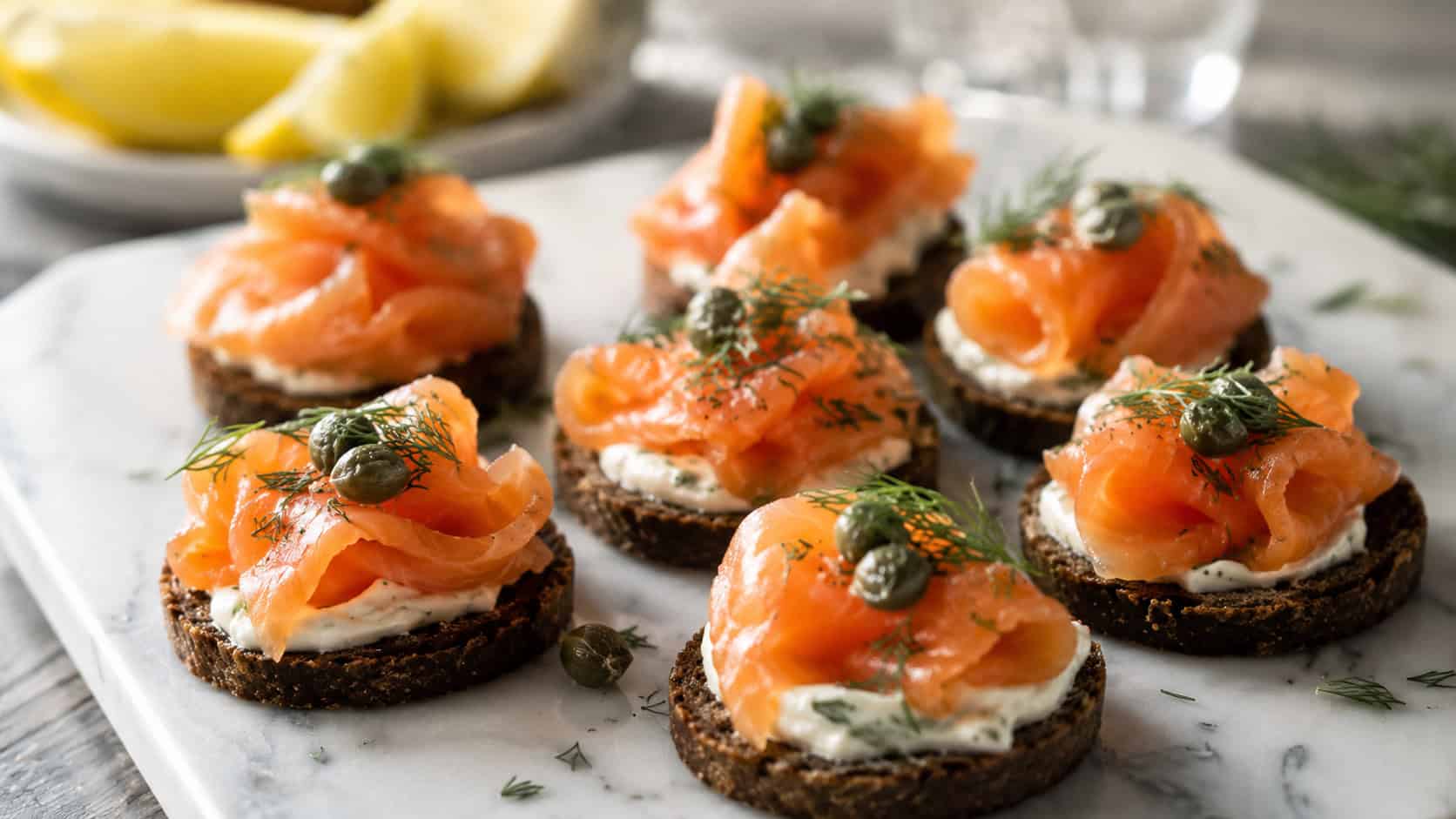 smoked salmon canapés arranged wide in landscape orientation on a white marble platter, dark rounds topped with pink salmon ribbons, dill, and capers, natural light, shallow depth of field, lemon wedges blurred in the background
