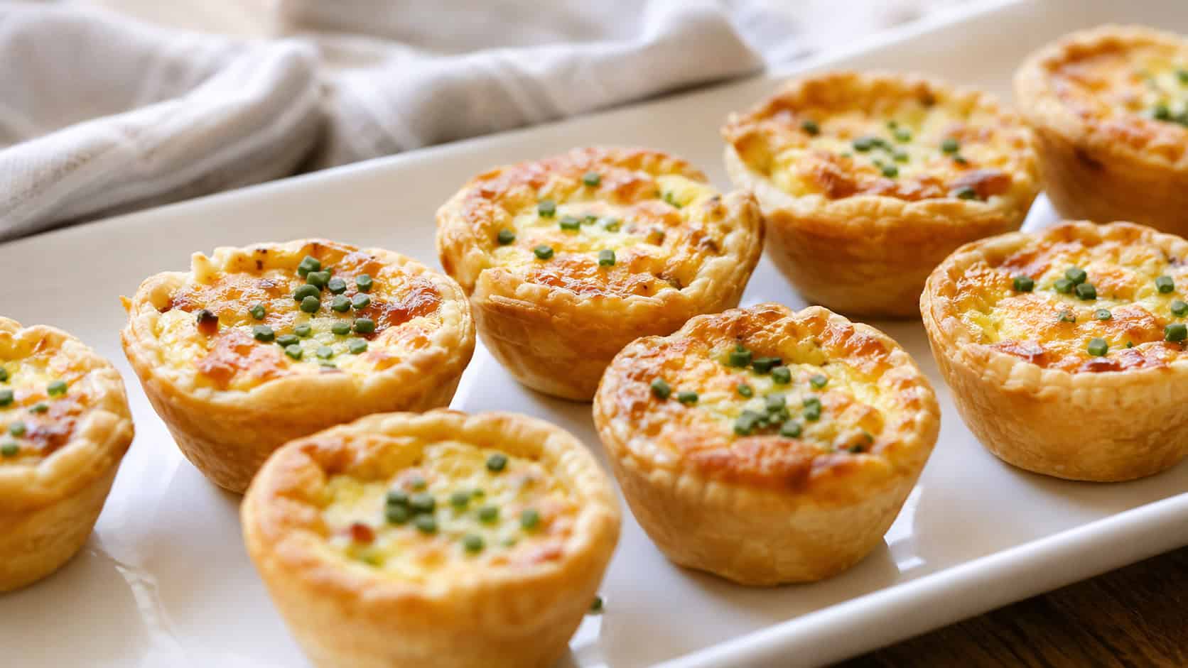 golden mini quiches arranged wide in landscape orientation on a white serving platter, flaky shells with puffed custard tops flecked with chives, natural light, shallow depth of field, a linen napkin blurred in the background