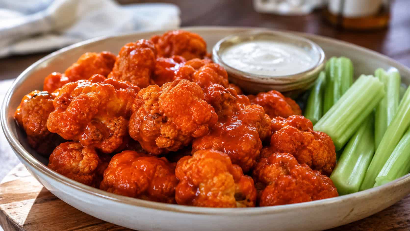 buffalo cauliflower bites glistening with red sauce piled in a wide shallow bowl in a horizontal shot, celery sticks and ranch dip beside them, warm natural light, shallow depth of field, landscape orientation