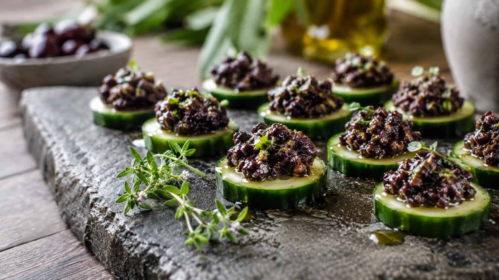 olive tapenade spooned over cucumber rounds on a stone platter in a wide horizontal shot, dark glossy olives, drizzle of oil, fresh herb sprig, natural light, shallow depth of field, landscape orientation