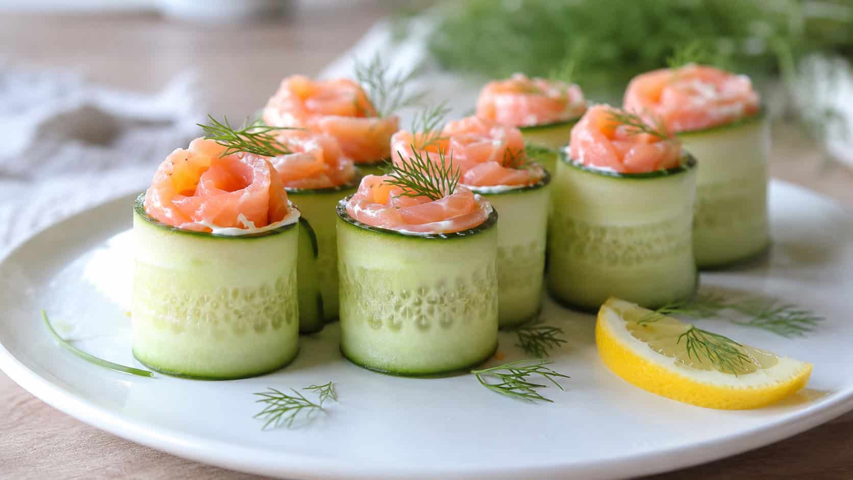 cucumber salmon rolls standing upright on a white plate in a wide horizontal shot, pink salmon spiral visible, dill sprigs, lemon wedge, bright airy natural light, shallow depth of field, landscape orientation