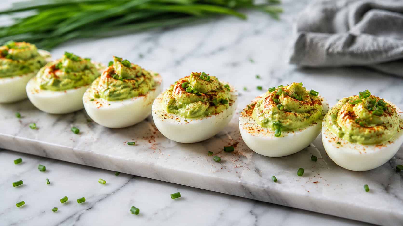 avocado deviled eggs lined up on a marble serving board in a wide horizontal shot, dusted with paprika, fresh chives scattered nearby, bright natural light, shallow depth of field, landscape orientation